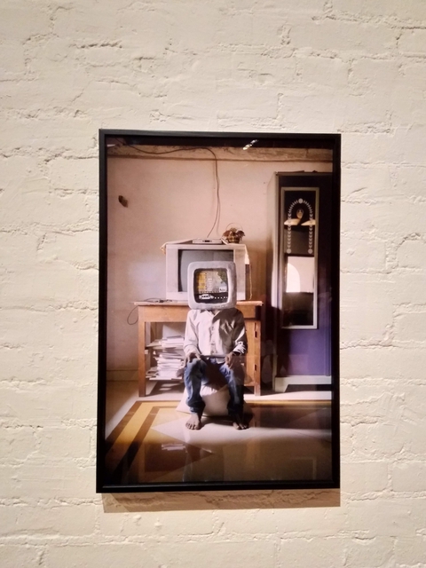 Photo of man wearing papier mache mask of a computer monitor, sitting in front of a CRT TV.